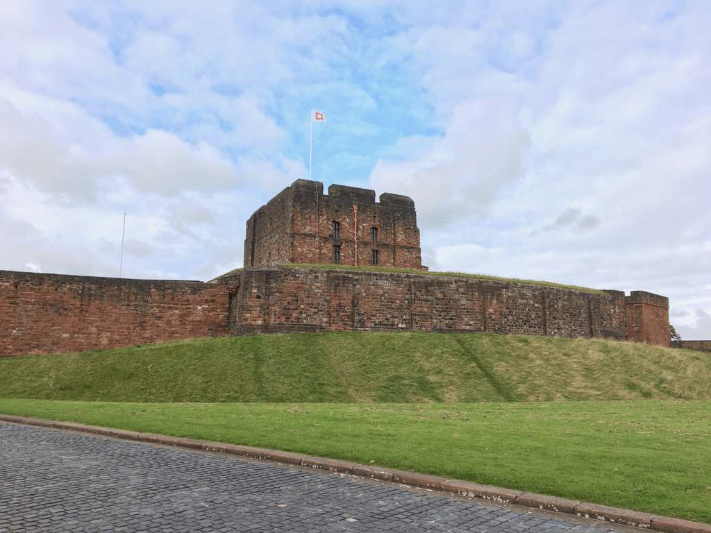 Carlisle Castle | Old stone castle with a flag on top, surrounded by grassy grounds and a cobblestone path in the foreground.