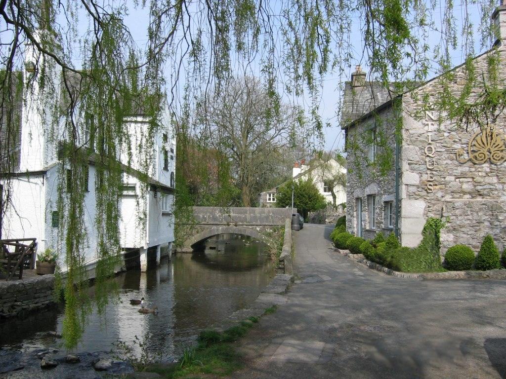 A quaint village scene with a stone bridge over a narrow stream, flanked by white and stone buildings, one marked "Antiques". Weeping willow branches drape over the scene and ducks swim in the water.