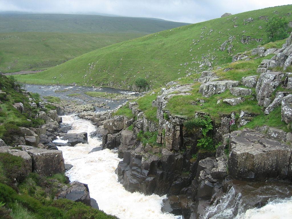 Waterfall cascading over rocks in a hilly landscape with green grass and sparse vegetation.