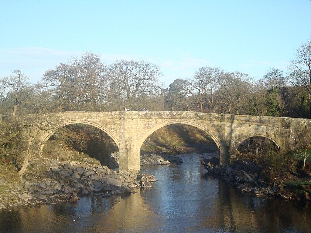 Stone bridge with three arches over a calm river, surrounded by trees.