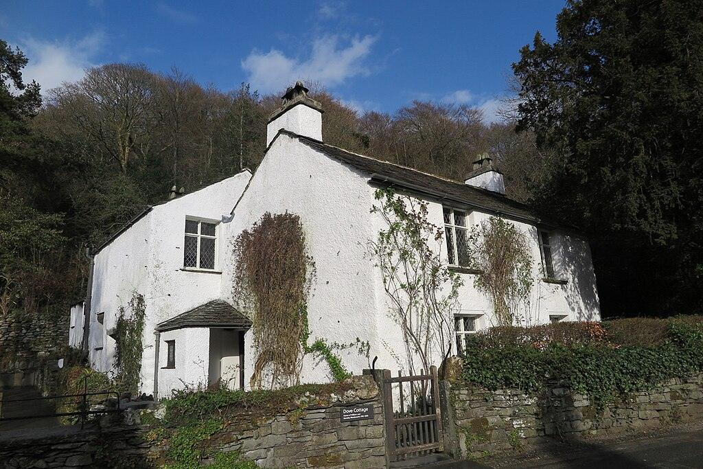 White cottage with ivy climbing up the front walls, surrounded by greenery and trees, under a blue sky with scattered clouds.