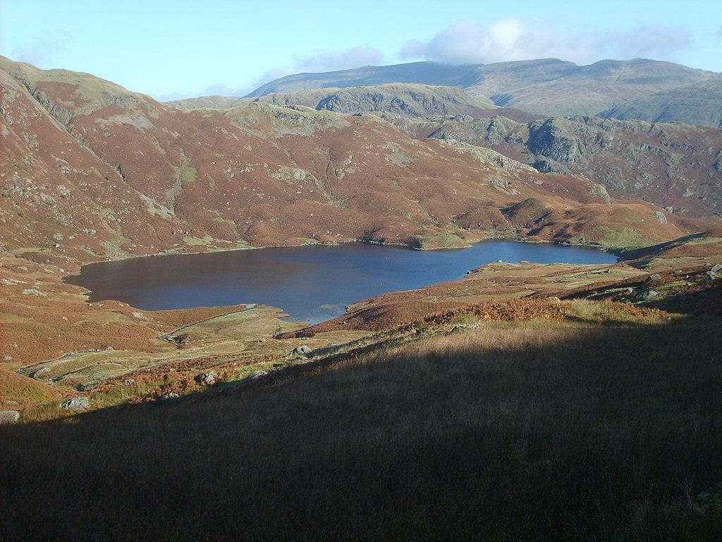 Mountainous landscape with a brownish lake surrounded by hills and a clear blue sky in the background.