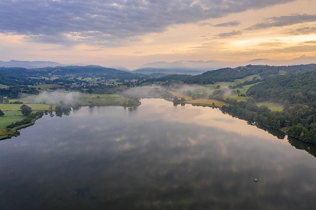 Aerial view of serene lake surrounded by green fields and forested hills, with mist rising and a cloudy sky at sunrise.