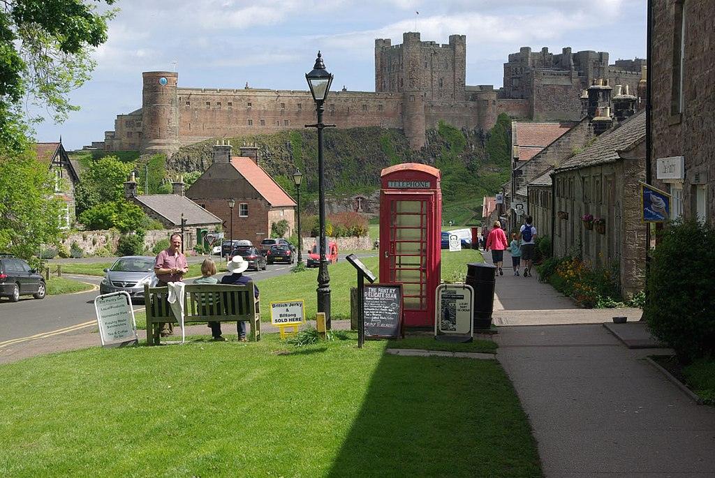 Bamburgh,A quaint village scene with a red British telephone box in the foreground, people sitting on benches, and a stone castle on a hill in the background.