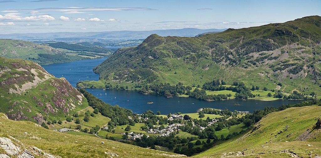 Glenridding, Cumbria | Aerial view of Ullswater in the Lake District, with a village, green hills, and boats on the water.