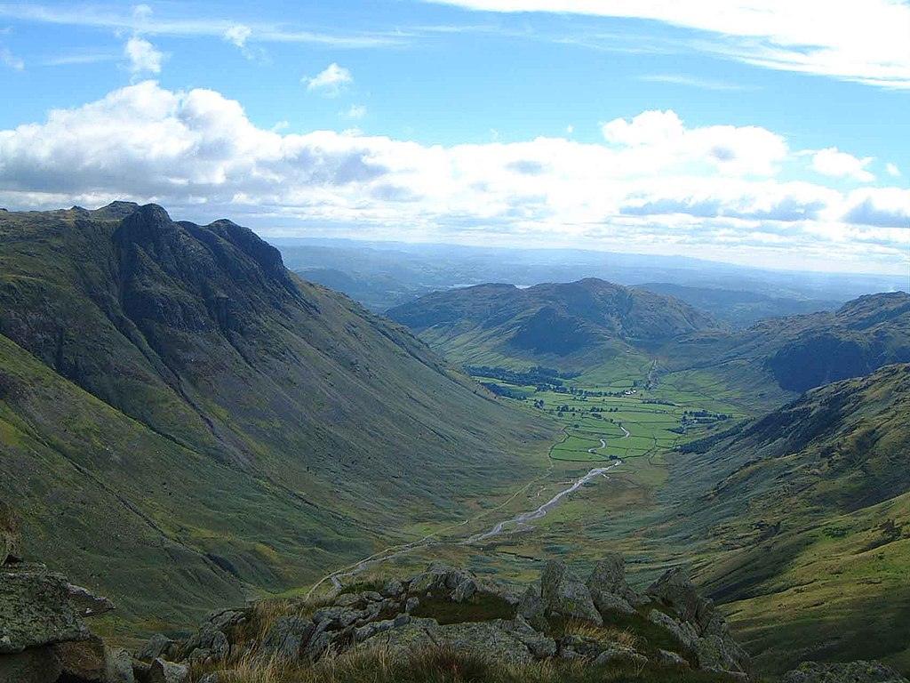 Scenic view of a lush valley from a high vantage point, featuring rugged, green hillsides and a winding stream, under a partly cloudy sky.