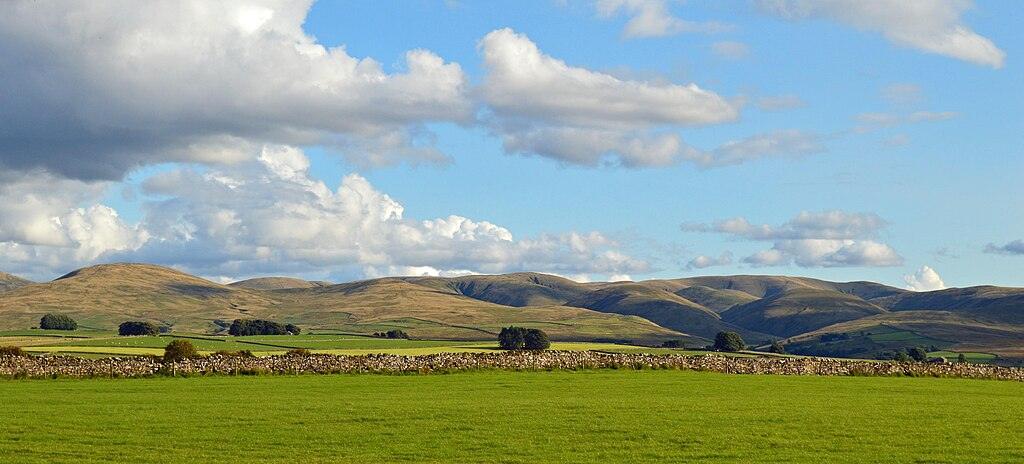 A scenic view of rolling hills under a partly cloudy sky, with a stone wall and green fields in the foreground.