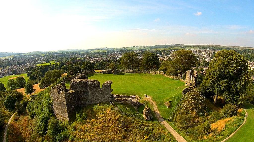 Aerial view of the ruins of Kendal Castle in Cumbria, England, surrounded by greenery with the town of kendal and countryside in the background.