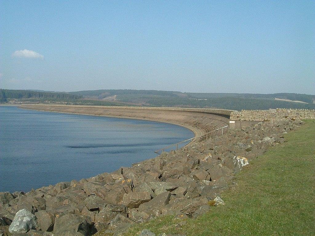 Curved dam structure with calm water on one side and rocky embankment on the other, under a clear blue sky.