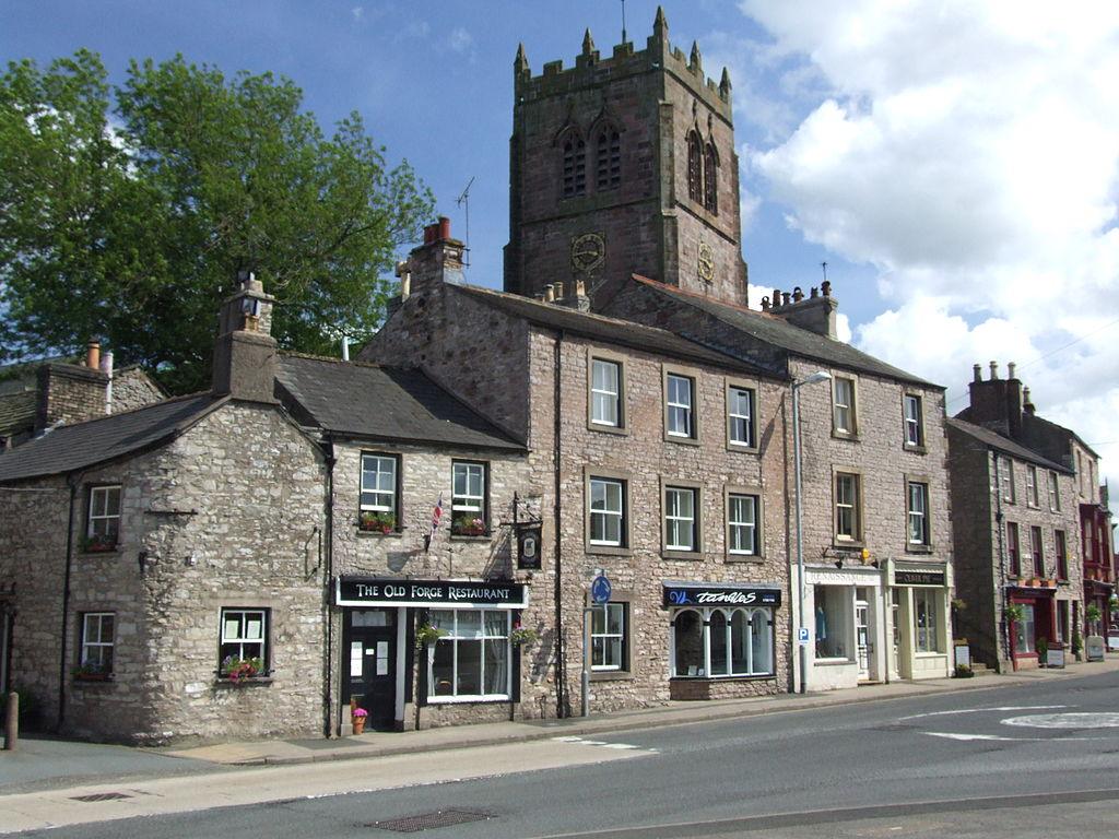 Kirkby Stephen, A Street scene in a small town with stone buildings, including "The Old Forge Restaurant" and a church with a tall tower in the background.