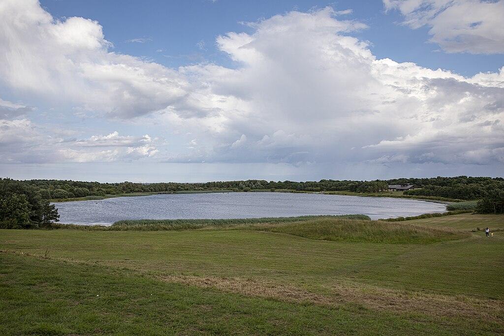 A serene lake surrounded by trees and grassy fields under a partly cloudy sky.