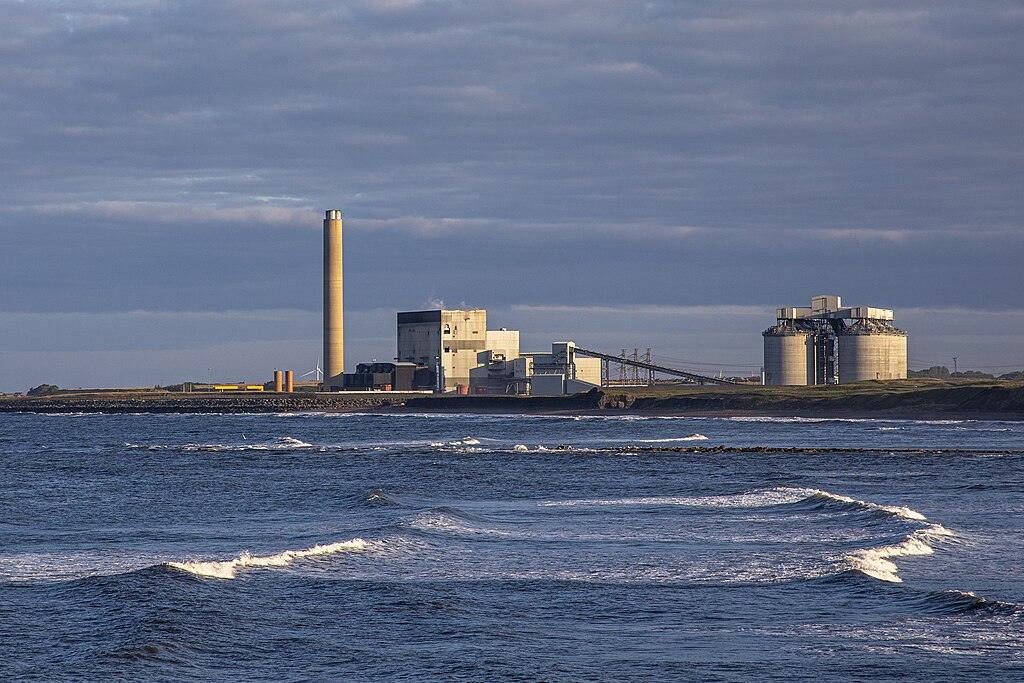 Industrial facility by the coastline with a tall smokestack and cylindrical storage tanks, partially illuminated by the setting sun, under a cloudy sky.