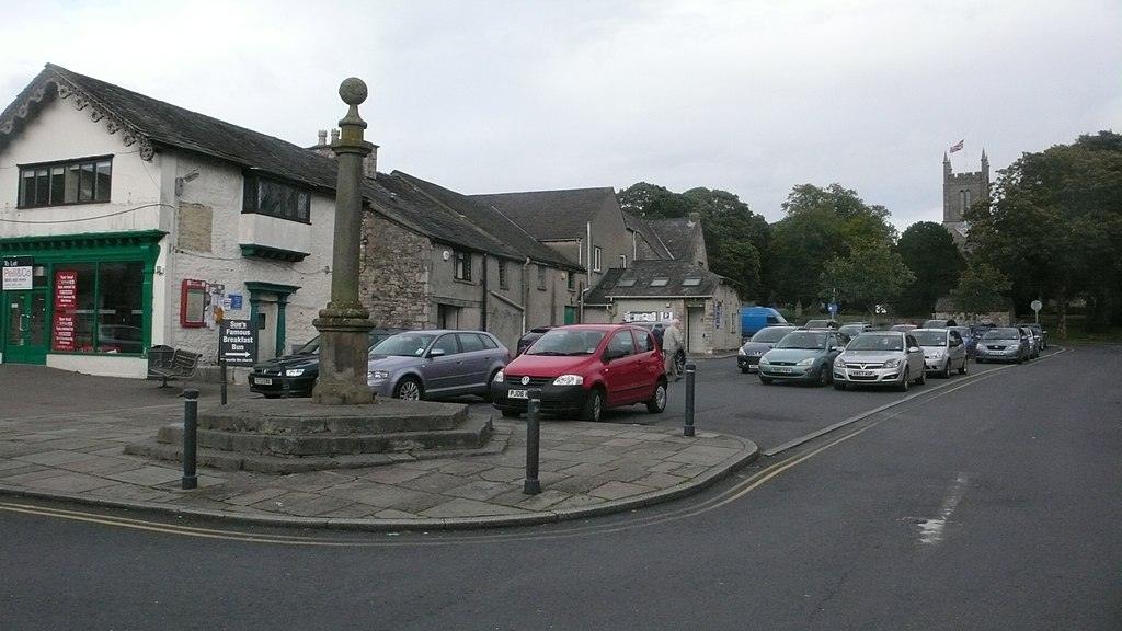 Market Square, Milnthorpe, A stone monument in a small town square, surrounded by parked cars and buildings; a church with a flag is visible in the background.