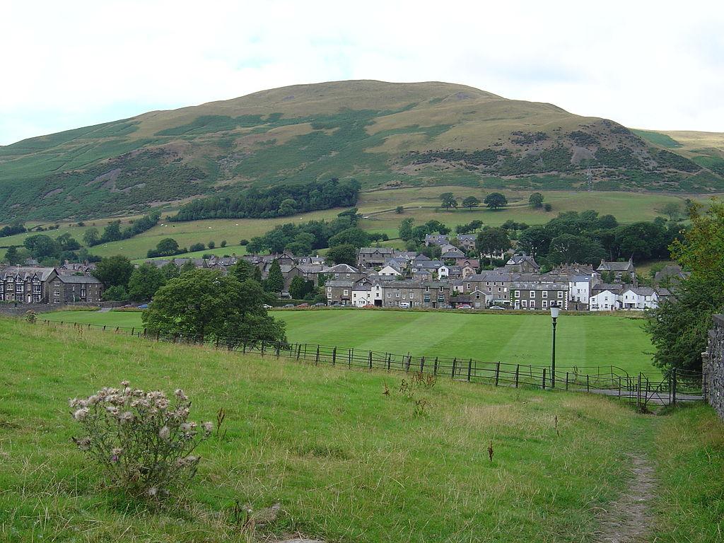 A rural village nestled at the foot of a green hill, with stone houses surrounded by fields and trees.