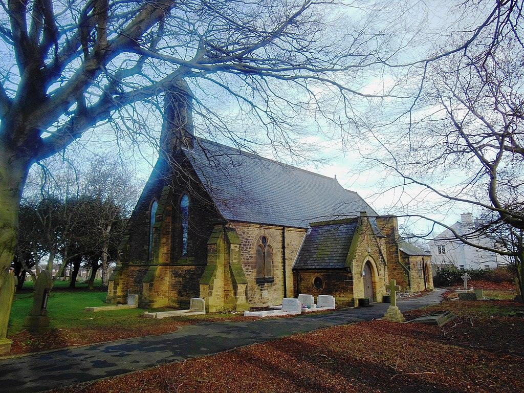 A stone church with gothic architecture, surrounded by leafless trees and gravestones, with a pathway leading to its entrance.