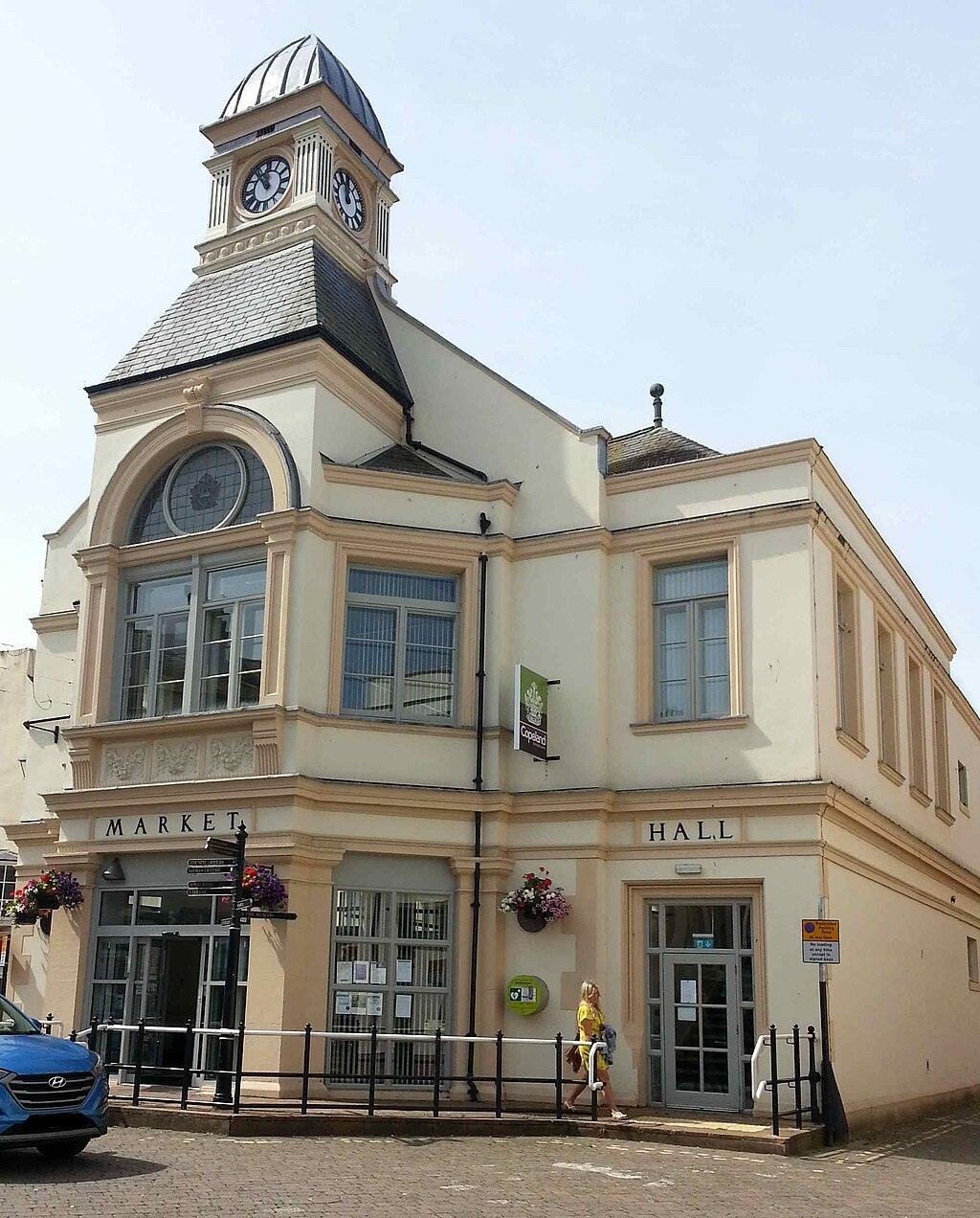 A historic market hall building with a clock tower, large windows, and hanging flower baskets, located in a paved area with a car parked in front.