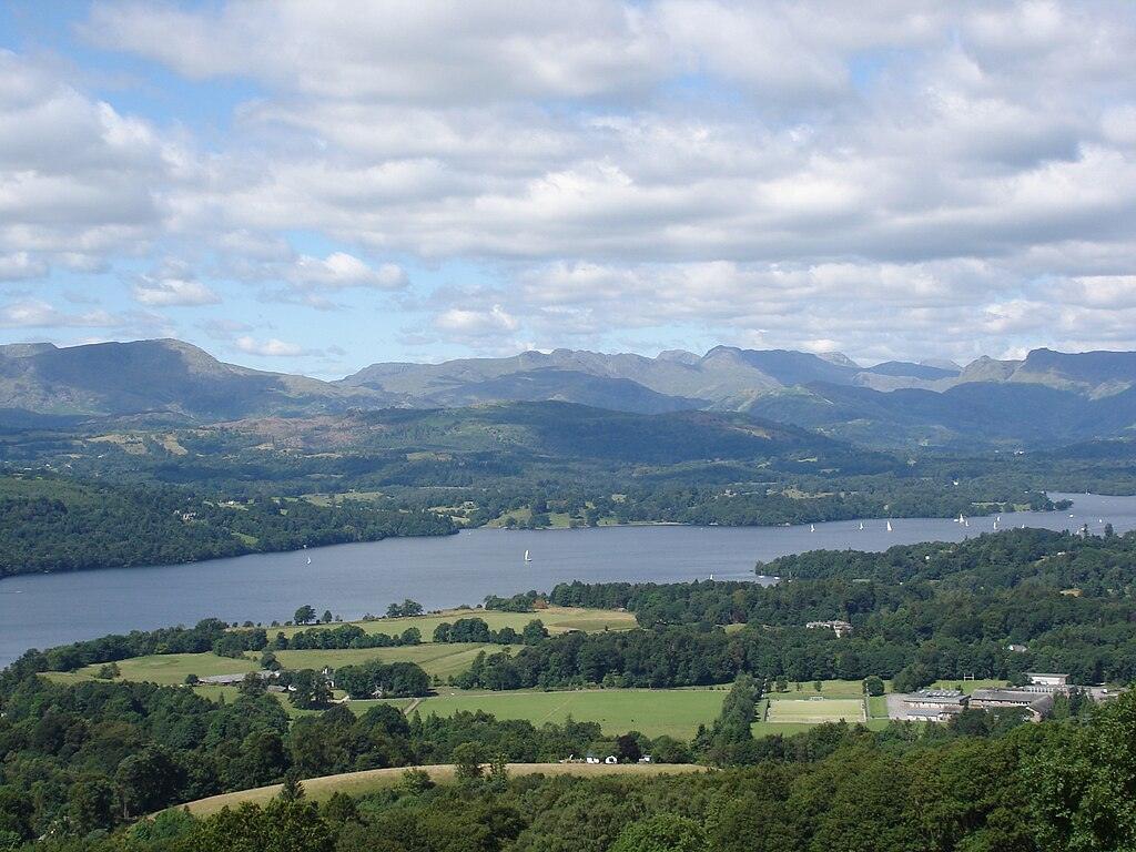 Panoramic view of Lake Windermere surrounded by wooded countryside and distant blue hills under a partly cloudy sky.