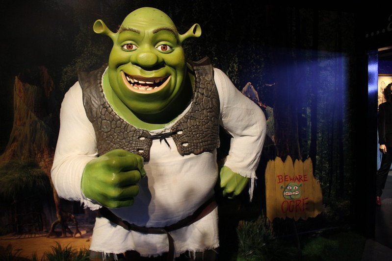 A wax figure of Shrek standing in a forest-themed setting, with a small sign reading "Beware Ogre" nearby.