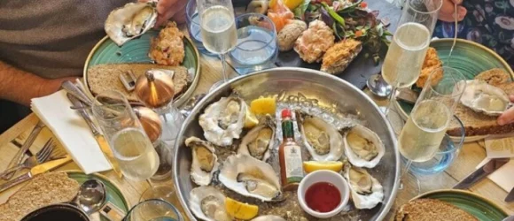 Seafood platter with oysters, lemon wedges, and a small bottle of Tabasco sauce on a table with champagne glasses, bread, and various dishes.