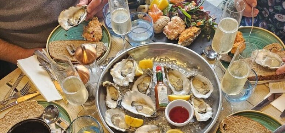 Seafood platter with oysters, lemon wedges, and a small bottle of Tabasco sauce on a table with champagne glasses, bread, and various dishes.