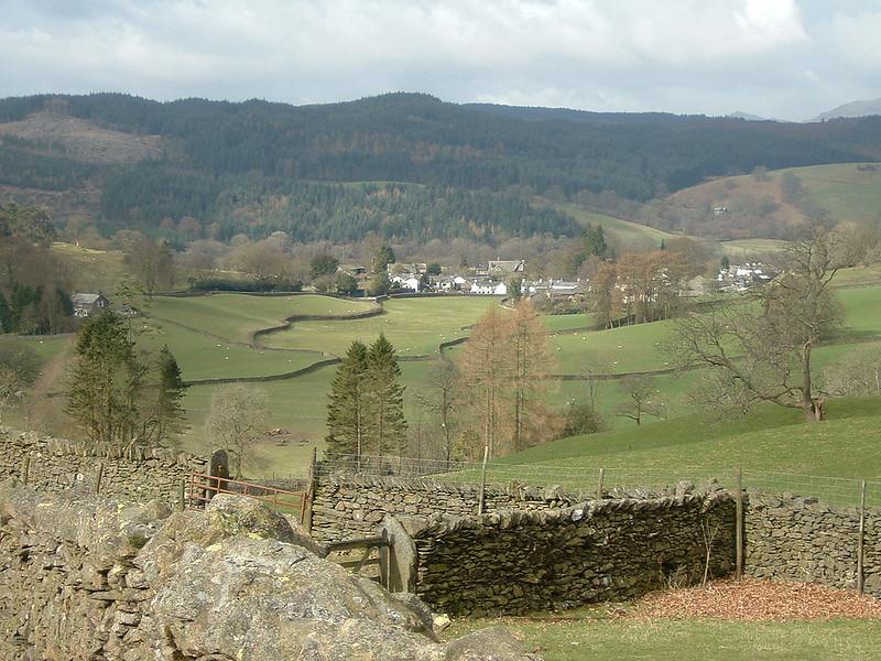 Scenic view of a rural landscape with green fields, stone walls, scattered trees, and a small village in the distance, set against a backdrop of forested hills.