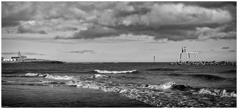 Black and white seascape with waves crashing on the shore, a distant structure on the left, and two people standing on a dock or pier on the right.