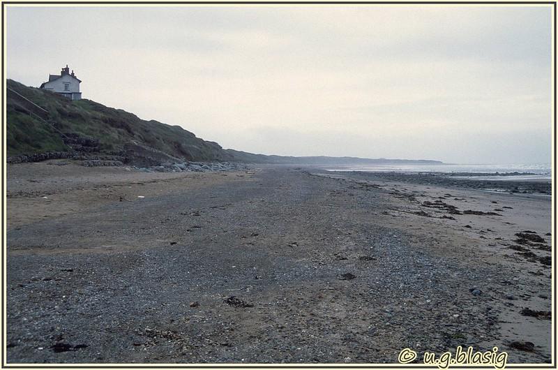 Seascale, Cumbria | A secluded beach with pebbles and sand, with gentle waves in the background and a solitary white house on a grassy hilltop to the left.