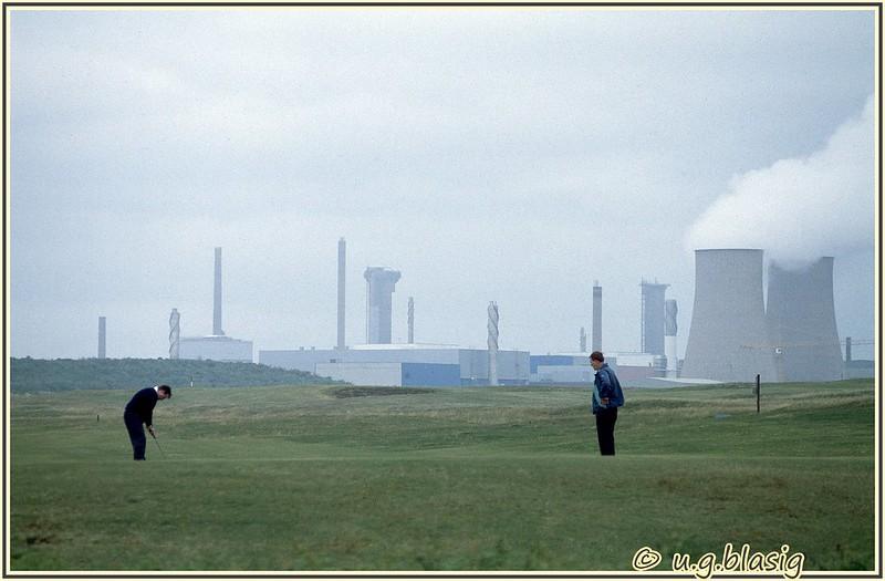 Two people playing golf on a grassy course with an industrial plant and smoke stacks in the background.