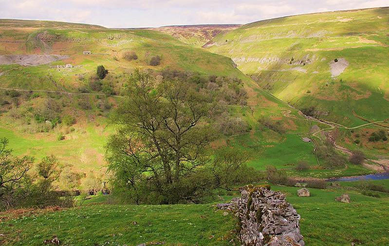 Lush green valley with rolling hills, scattered trees, and a meandering stream; some ruins visible on the hillside under a partly cloudy sky.