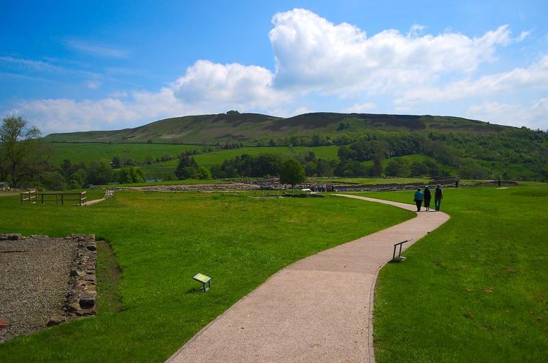 Vindolanda | People walk along a winding path through a grassy field with hills in the background under a partly cloudy sky.