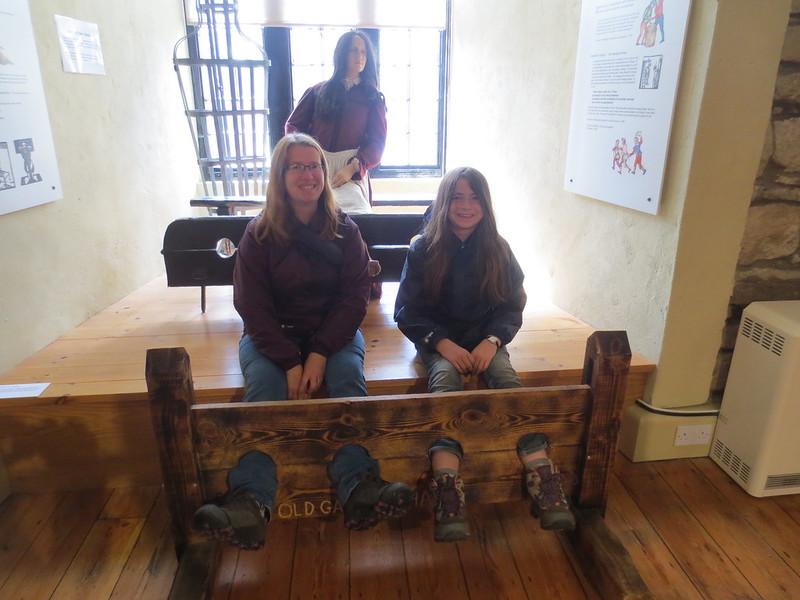 Two people sitting with their feet in wooden stocks, with a mannequin in historical clothing behind them in a museum setting.