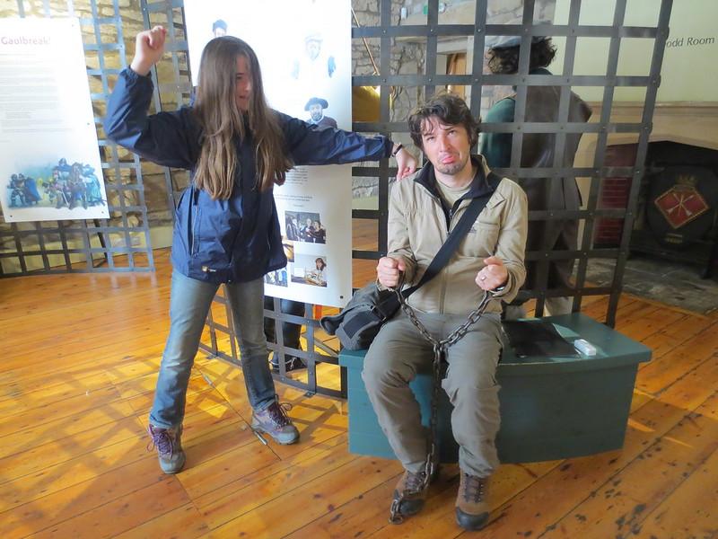 A girl playfully pretends to arrest a man sitting in shackles inside a museum exhibit.