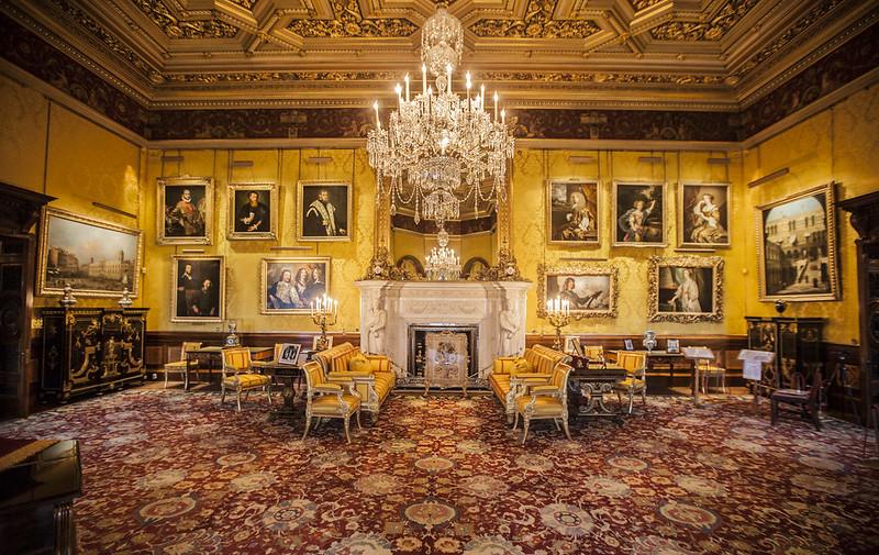 Ornate room with yellow walls, elaborate ceiling, chandelier, large framed paintings, luxurious chairs, and a fireplace with a marble mantle.