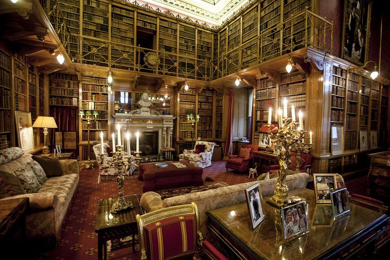 Luxurious library with tall, ornate bookshelves filled with books, a fireplace, plush seating, and a candelabrum on a table decorated with framed photographs.