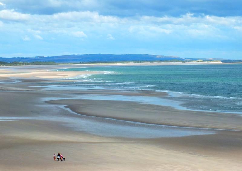 A wide sandy beach with shallow pools of water, a small group of people in the foreground, and distant hills under a cloudy sky.