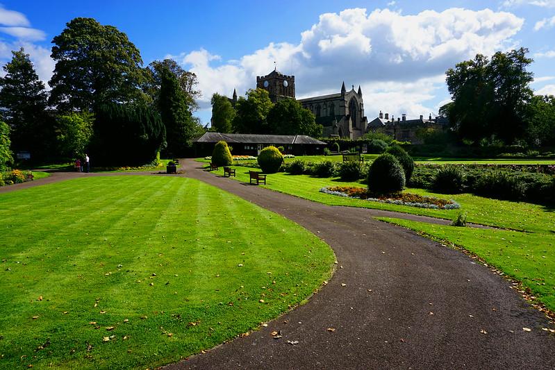 Hexham Abbey Gardens | A landscaped park with a curved path leading to benches and flowerbeds, with a large historic building and trees in the background.