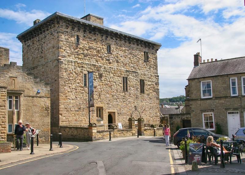 A stone building with small windows in a quaint town, with people walking and sitting outside at a café.