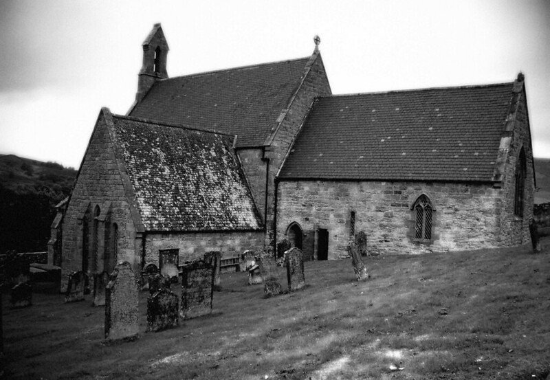 A black and white photograph of an old stone church with a pitched roof, surrounded by a graveyard with several tombstones on a grassy slope.