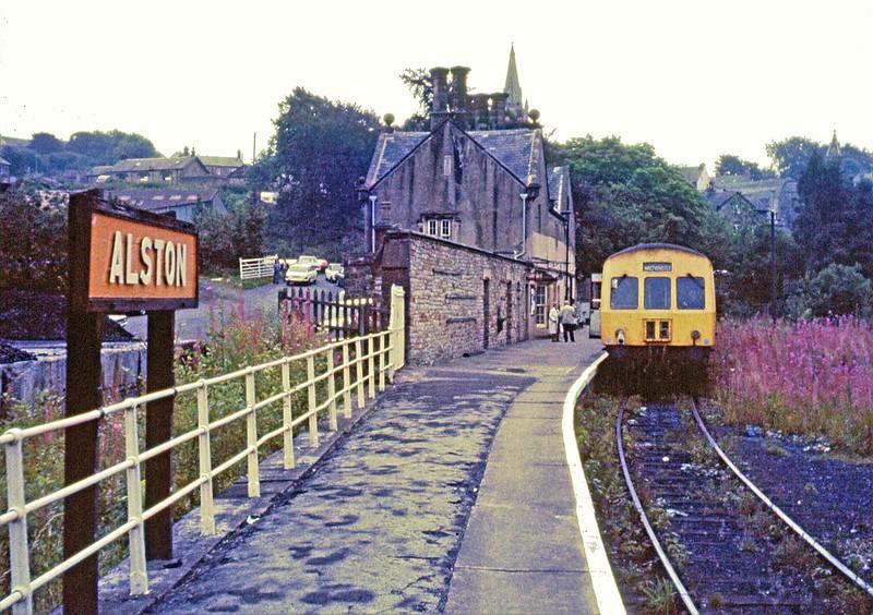 Alston railway station with a train at the platform, buildings and houses in the background.