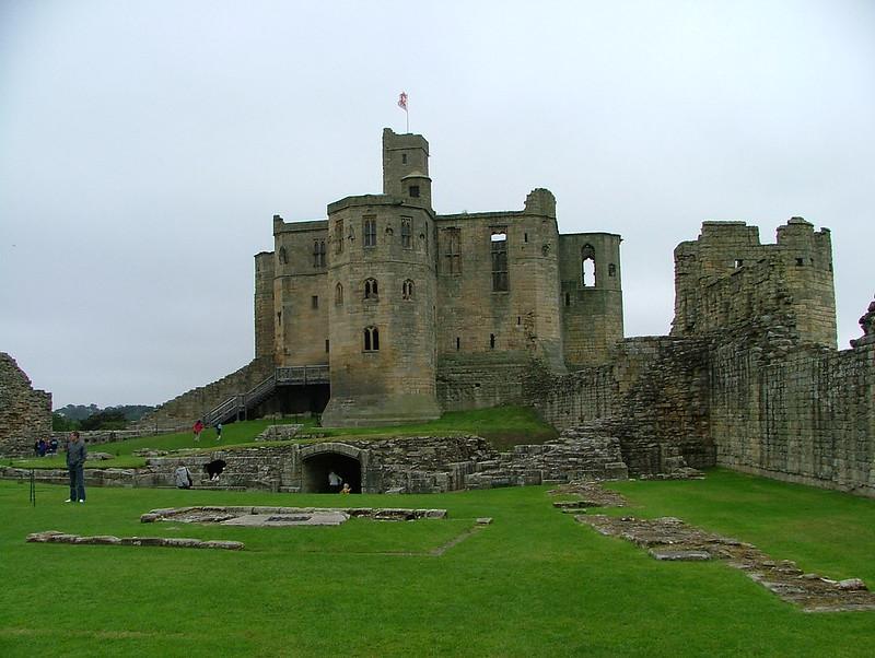 Warkworth Castle, Northumberland | A medieval stone castle with a central keep and surrounding ruins, situated on a grassy landscape under a cloudy sky.