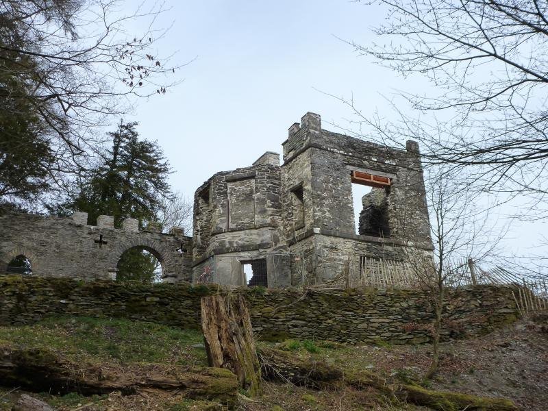 Ruins of a stone building with an arched wall next to it, surrounded by trees and greenery.