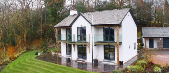 Two-storey modern house with white walls, large glass windows, and balconies, surrounded by a well-maintained garden and lawn, with a backdrop of trees.