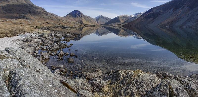 Panoramic view of a calm lake reflecting mountains and a clear sky, surrounded by rocky terrain and gentle slopes.