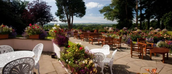 Outdoor garden with white metal chairs, wooden benches, and tables surrounded by colourful flowers and trees, overlooking a scenic landscape.