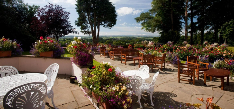 Outdoor garden with white metal chairs, wooden benches, and tables surrounded by colourful flowers and trees, overlooking a scenic landscape.