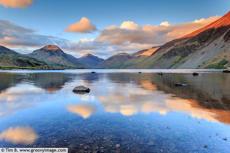 Lake District, A Peaceful mountain lake at sunset, with reflections of surrounding hills and a partly cloudy sky on the calm water.