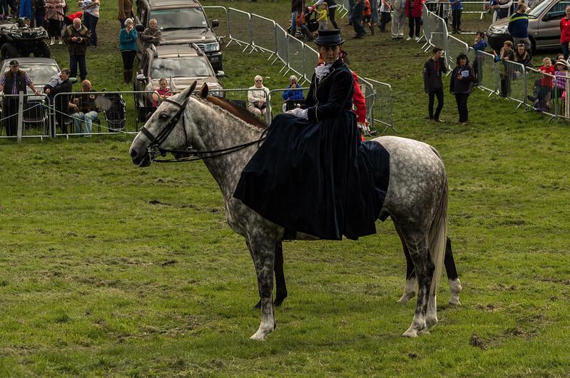 A person dressed in formal attire, including a long coat and hat, riding a grey horse in a grassy field with spectators and parked cars in the background.