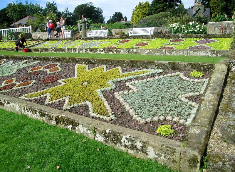 Ornate flowerbeds with various colourful patterns surrounded by a stone border, people and benches visible in the background.