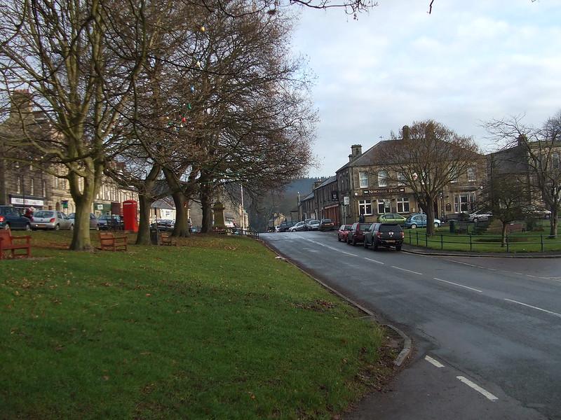 Rothbury a small town street with parked cars alongside a green space with bare trees, benches, and a red telephone box, and stone buildings in the background.