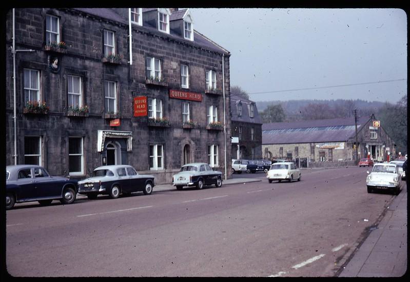 A street with parked vintage cars in front of a stone building named "Queens Head," featuring flower boxes on the windows.